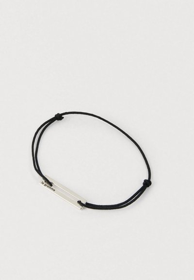 Fekete állítható zsinór karkötő, vékony vízszintes ezüst rúddal, amelyre a "le gramme" felirat van gravírozva, letisztult világos háttér előtt.