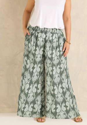 Kvinde iført grønne bukser med vide ben og hvide paisley-mønstre, hvid ærmeløs top, sandaler og armbånd, stående med hænderne i lommerne.