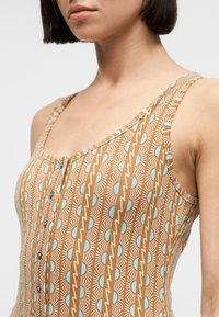 Γυναίκα που φοράει αμάνικο καφέ φόρεμα με μπλε ημικύκλιο και κίτρινο ζιγκ ζαγκ σχέδιο, με κουμπιά μπροστά και ίσιο καρέ κούρεμα.