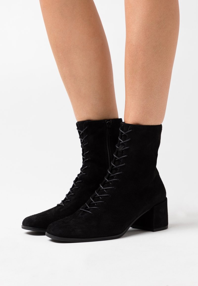Bottines en daim noir à lacets avec talons blocs portées sur des jambes nues sur fond blanc.