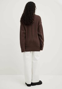 Brauner oversized Kabelstrick-Pullover mit geripptem Saum und Ärmeln, kombiniert mit weißen, locker sitzenden Hosen und schwarzen klobigen Stiefeln.