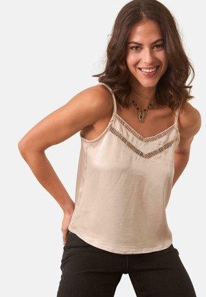 Camisola de satén beige con escote en V poco profundo adornado con cuentas y tirantes finos. Combinada con jeans oscuros, el modelo posa con las manos en las caderas.