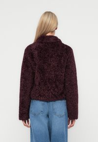 Bordeaux nepbontjack met lange mouwen, een kraag en een gestructureerde afwerking, gecombineerd met blauwe wijdvallende jeans. Achteraanzicht weergegeven.
