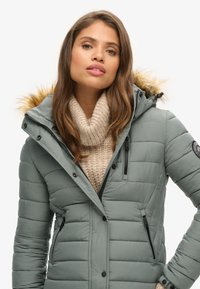 Chaqueta acolchada verde claro con capucha de piel sintética, bolsillos con cremallera y un cuello alto, llevada sobre un suéter de punto grueso color crema.