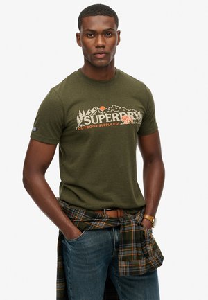 Olijfgroene katoenen T-shirt met een bedrukte graphic met de tekst "SUPERDRY" en outdoor-thema beelden. Gekozen met een geruite blouse om de taille geknoopt.