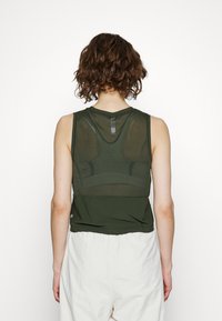 Top sem mangas verde-oliva com um painel traseiro transparente, decote arredondado e parte inferior texturizada, combinado com calças largas de cor clara.
