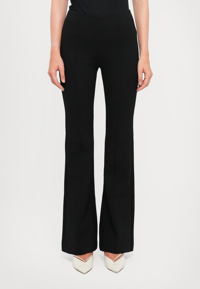 Pantalon noir à jambes larges avec une texture lisse et une taille ajustée, présentant des coutures avant et une forme longue et fluide. Porté avec des talons à bouts pointus.
