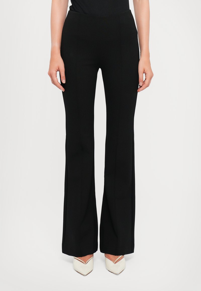 Black flared trousers with a smooth texture, high waist, and vertical seam details; paired with white pointed-toe heels.