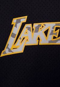 Tela de malla negra con la palabra "LAKERS" en amarillo negrita y acentos de camuflaje gris. La textura notable incluye agujeros de ventilación en la tela.