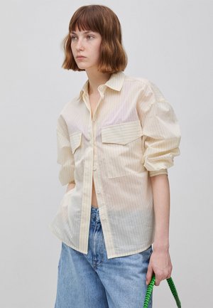 Jonge vrouw met kort bruin haar, gekleed in een licht gestreept overhemd met knopen en een blauwe spijkerbroek, met een handvat van een groene gevlochten tas in haar hand.