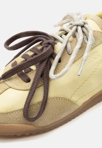 Κίτρινο δερμάτινο αθλητικό παπούτσι με μπεζ σουέτ λεπτομέρειες, που διαθέτει δύο ζευγάρια δεμένων κορδονιών σε καφέ και ανοιχτό γκρι.