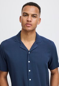 Camicia blu navy a maniche corte con colletto a punta, texture liscia e sei bottoni bianchi sul davanti. Nessun motivo o dettaglio.
