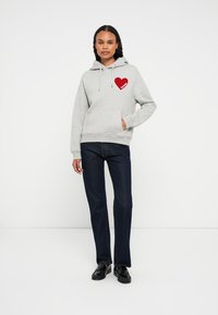 Kvinne iført grå hettegenser med rødt hjerte og teksten "FUTURE", mørkejeans og svarte loafers, står mot en enkel lys bakgrunn.