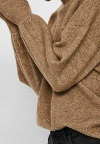 Pull en maille marron avec une finition texturée, manches longues et coupe décontractée, porté avec un pantalon noir.