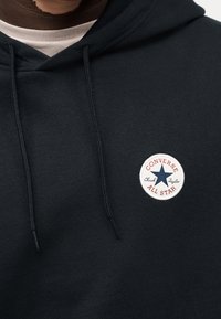 Μαύρο φούτερ με κουκούλα και κορδόνι, με στρογγυλό λευκό έμβλημα που αναγράφει «Converse All Star» και μπλε αστέρι. Μαλακή υφή, casual σχέδιο.