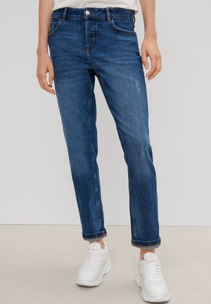 Dunkelblaue Denim-Jeans mit geradem Schnitt, aufgerollten Säumen, Fronttaschen und einem kupferfarbenen Knopf. Kombiniert mit weißen Turnschuhen.
