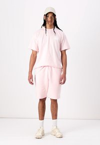 Helles rosa Baumwoll-T-Shirt und passende Shorts mit lockerer Passform. Mit geripptem Kragen, Seitentaschen und kleinen Logo-Details. Weiße Sneaker und Kappe.