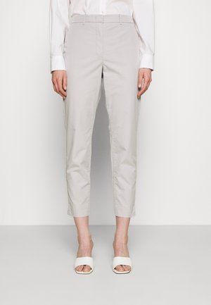 Vrouw gekleed in lichtbeige cropped broek, wit geknoopt overhemd en witte open-toe hakken, staand tegen een effen witte achtergrond.