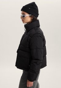 Giacca a vento nera imbottita con colletto alto, tasca frontale e chiusura con bottoni. Indossata con un berretto lavorato a maglia aderente e occhiali da sole trasparenti.