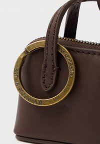 Geantă maro din piele cu un accent de inel metalic auriu, având un mâner cusut și un detaliu cu fermoar de-a lungul marginii superioare.