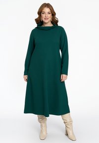 Langes grünes Kleid mit Kapuze und langen Ärmeln, locker geschnitten und mit glatter Textur. Kombiniert mit beigen, lässigen kniehohen Stiefeln.
