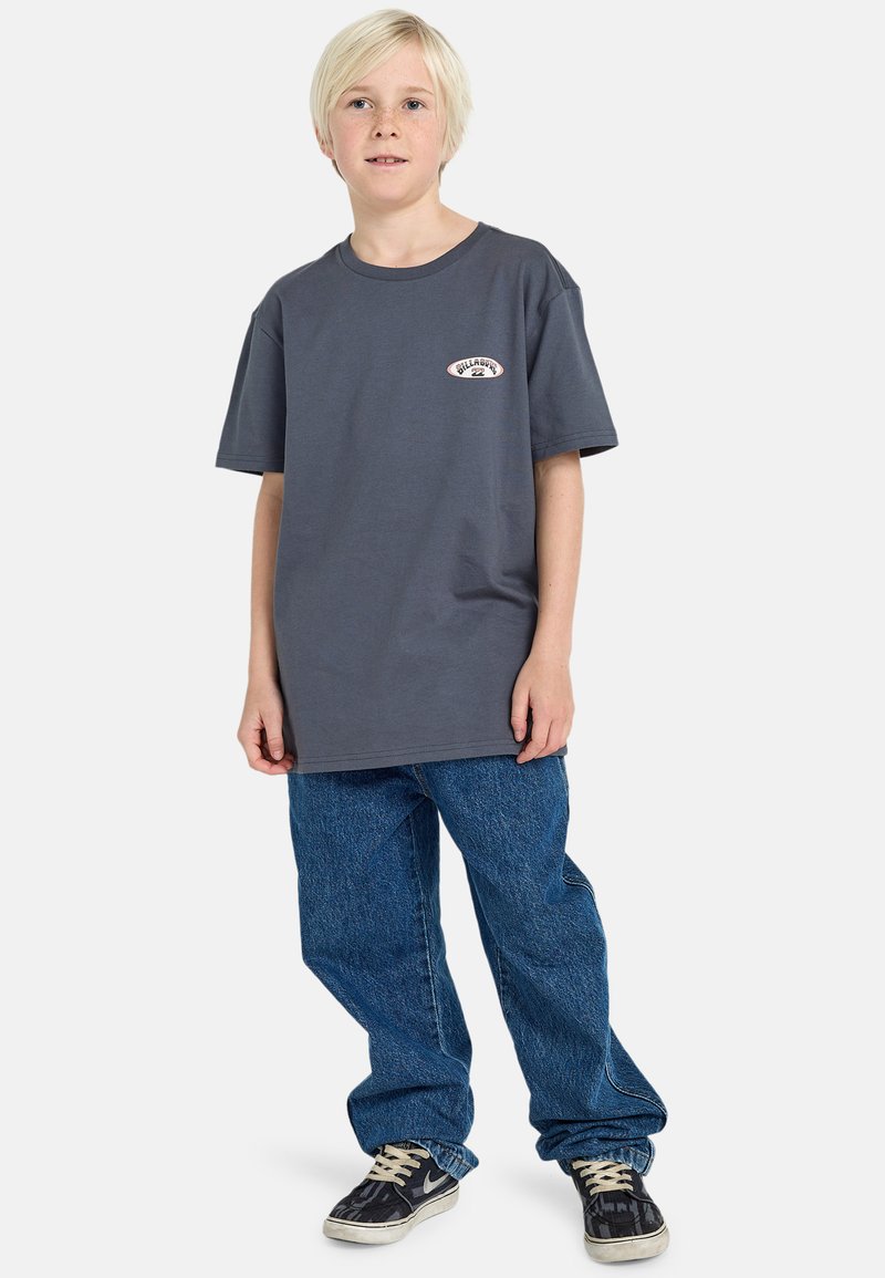 T-shirt en coton gris avec un col rond et un patch logo, associé à un jean en denim bleu ample et des sneakers noires avec des semelles blanches.