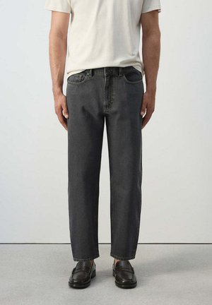 Hombre con vaqueros negros de corte holgado, camisa blanca de manga corta y mocasines de cuero marrón oscuro de pie sobre un suelo gris.