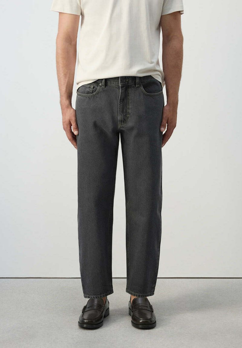 Homme portant un jean noir ample, une chemise blanche à manches courtes et des mocassins en cuir marron foncé debout sur un sol gris.
