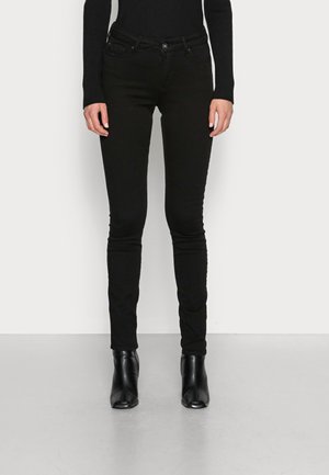 Person trägt eng anliegende schwarze Jeans, schwarzes Langarmoberteil und schwarze Stiefeletten, steht vor einem einfarbigen hellen Hintergrund.