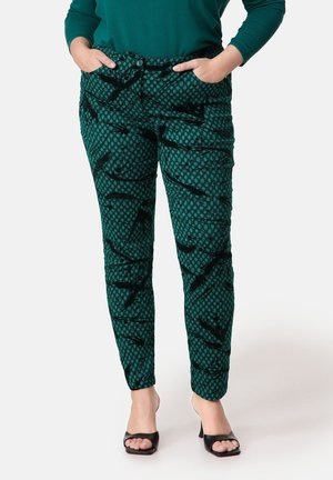 Vrouw draagt groene slim-fit broek met patroon, handen in de zakken en zwarte open-toe hak sandalen, staand op een witte achtergrond.