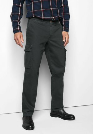Mann trägt dunkelgrüne Cargohosen, schwarze Lederschuhe und ein marineblau kariertes Hemd und steht an einer weißen Wand.