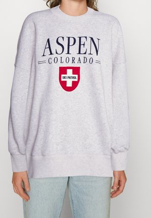 Lysegrå sweatshirt med teksten "ASPEN COLORADO" og et rødt ski-patrulje logo, båret med lyseblå jeans.