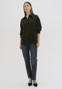 Mørkegrøn skjorte med knapper og krave, løs pasform og oprullede ærmer, parret med lyse vaskede jeans og sorte spidse hæle.
