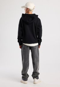 Felpa nera con cappuccio, caratterizzata da una texture morbida e una tasca a marsupio. Indossata con jeans grigi larghi e sneaker bianche, vista da dietro.