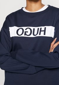 Sudadera azul marino con una franja horizontal blanca que presenta el texto "HUGO" en letras negras negritas; tejido suave y texturizado con puños acanalados.