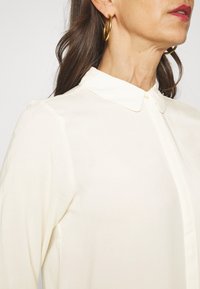 Blusa color crema con colletto classico, chiusura frontale a bottoni e tessuto morbido e liscio. Presenta maniche arrotolate e una vestibilità su misura.