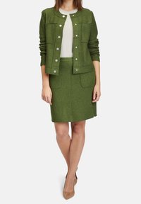 Green wool blend jacket with metallic buttons, worn over a light top and paired with a matching pencil skirt, featuring front pockets.