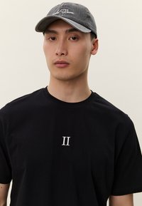 Melna kokvilnas T-krekls ar baltu "II" apdruku priekšpusē, kopā ar pelēku cepuri, uzšūtu ar "La Deuce". Vienkāršs un tīrs dizains.