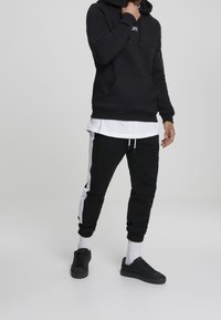 Svart hoodie med framficka och dragsko; lager-på-lager med en vit skjorta. Svarta sweatpants med vita sidostradar. Svarta sneakers och vita strumpor.