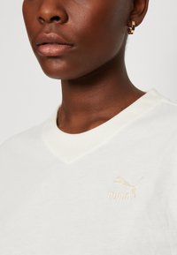 T-shirt bianco in cotone con scollo a V che presenta un discreto logo Puma ricamato in oro sul lato sinistro del petto e una texture liscia e morbida.