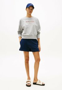 Felpa grigia con polsini a coste, pantaloni corti navy e sandali beige con suola nera. Cappellino da baseball in blu scuro. Dettagli del logo semplici.