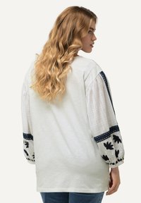 Blusa blanca con mangas largas texturizadas que presentan bordados florales en azul marino y acentos de lunares. Corte casual con escote redondo y fondo dobladillo.