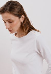 Camicia bianca a maniche lunghe con collo rotondo e dettagli in bottone dorato sulle spalle. Tessuto di cotone morbido con una finitura liscia.