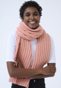 Écharpe tricotée rose à côtes en matériau doux, enroulée autour du cou. Le modèle porte un haut blanc et un pantalon noir, les bras croisés.