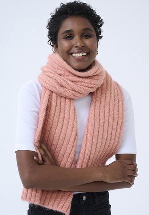 Sciarpa a coste rosa realizzata in morbido materiale, drappeggiata attorno al collo. Il modello indossa una maglietta bianca e pantaloni neri, con le braccia incrociate.