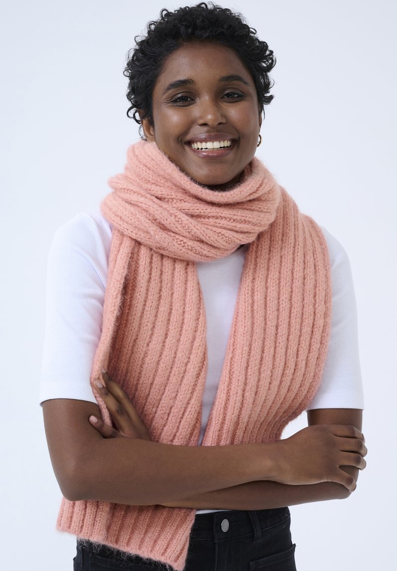Écharpe tricotée rose à côtes en matériau doux, enroulée autour du cou. Le modèle porte un haut blanc et un pantalon noir, les bras croisés.