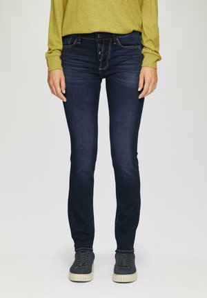 Dunkelblaue Skinny-Jeans mit glatter Textur, ausgestattet mit drei Knöpfen am Bund und kontrastierendem Nähen. Kombiniert mit grauen Turnschuhen.