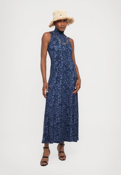 Rochie maxi florală albastră marină, cu guler înalt și fustă plisată. Modelul poartă sandale negre cu bretele și o pălărie din paie cu margine desfăcută.