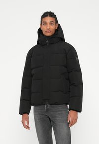 Černá zateplená bunda s kapucí, má přední zip a hladkou texturu. Model má na sobě šedé džíny, což ukazuje na ležérní vzhled.