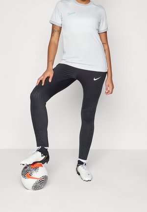Persona con mallas negras de Nike y tacos blancos, apoyando el pie sobre un balón de fútbol blanco y negro con el logo naranja de Nike.